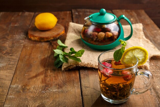 Zielona herbata w szklanej filiżance z truskawkami miętą i cytryną na drewnianym stole oraz czajniczkiem i cytryną w talerzu i listkami mięty