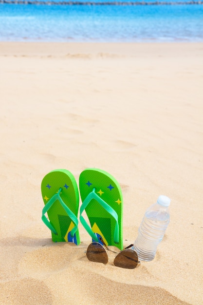 Zielone sandały na piaszczystej plaży z butelką czystej wody i okularami