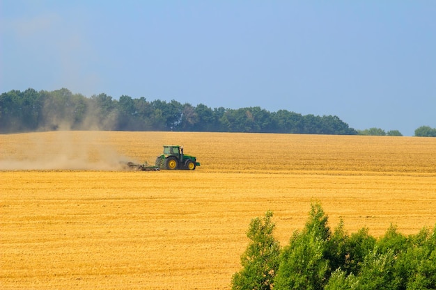 Zielony traktor z broną pracuje na żółtym polu jesienią