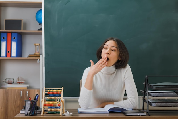 ziewanie z zamkniętymi oczami młoda nauczycielka siedzi przy biurku z włączonymi narzędziami szkolnymi w klasie