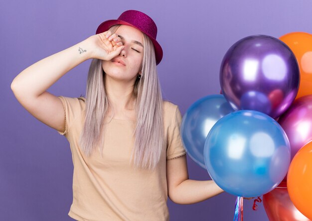 Ziewanie z zamkniętymi oczami młoda piękna dziewczyna w imprezowym kapeluszu trzymająca balony ocierająca oko ręką odizolowaną na niebieskiej ścianie