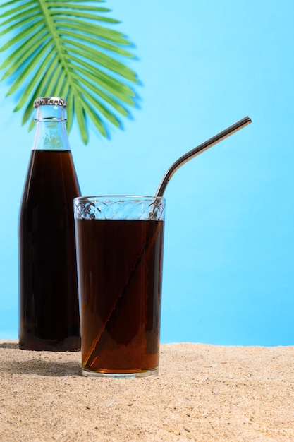 Zimny napój w szklance i butelce na piasku na niebieskim tle z liściem palmowym