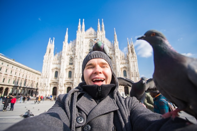 Zimowe podróże, wakacje i koncepcja ptaków - przystojny mężczyzna turysta ze śmiesznymi gołębiami robi zdjęcie selfie przed słynną katedrą Duomo w Mediolanie.