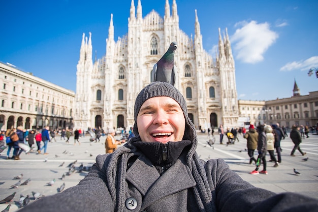 Zimowe podróże, wakacje i koncepcja ptaków - przystojny mężczyzna turysta ze śmiesznymi gołębiami robi zdjęcie selfie przed słynną katedrą Duomo w Mediolanie.