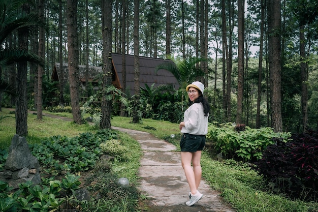 Zimowe podróże zrelaksuj się na wakacjachPortret Azjatycka turystka w białej sukni z kapeluszem stoi w sosnowej kabinie w sosnowym lesie na szlaku przyrodniczym w parku leśnym Doi Bo LuangChiang Mai Tajlandia