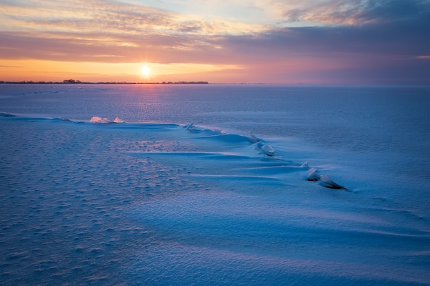 Zimowy krajobraz z zamarzniętym jeziorem, pęknięciami i zachodem słońca na niebie.