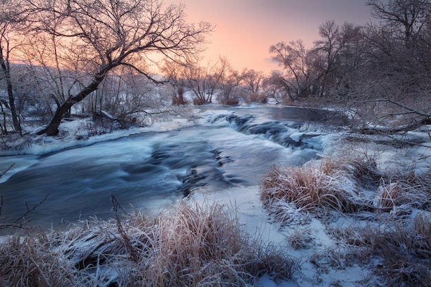 Zimowy pejzaż z ośnieżonymi drzewami, piękna zamarznięta rzeka o zachodzie słońca