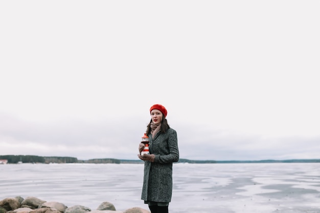 Zimowy portret młodej kobiety w płaszczu i czerwonym kapeluszu, trzymając ozdobną latarnię morską i stojąc na brzegu zamarzniętego morza. zima, podróż, morze w tle. wietrzna pogoda, niesamowite lodowe wybrzeże