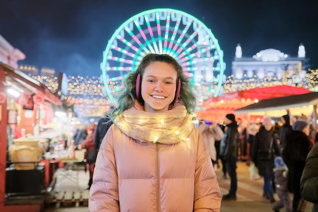 Zimowy portret szczęśliwa młoda nastolatka kobiece na jarmark bożonarodzeniowy. Światła, jasne girlandy, tło diabelskiego młyna