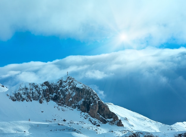 Zimowy stok górski z trasą narciarską i krzyżem na szczycie skały (Austria).