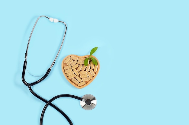 Ziołowe tabletki w drewnianej płytce w kształcie serca i stetoskop na niebieskim tle Medycyna alternatywna