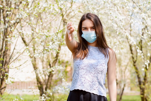 Zirytowana dziewczyna stojąca w kwitnącym parku pokazująca kciuki w dół, aby pokazać swoje podrażnienie i niechęć do ciągłego noszenia maski medycznej, aby zapobiec rozprzestrzenianiu się koronawirusa