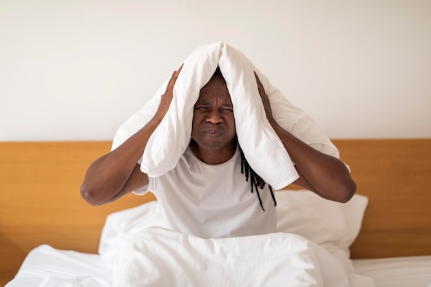 Zirytowany czarny facet zakrywający uszy poduszką podczas siedzenia w łóżku