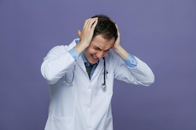Zirytowany młody lekarz mężczyzna ubrany w szatę medyczną i stetoskop wokół szyi, zginając głowę w dół, zamykając uszy rękami z zamkniętymi oczami na fioletowym tle