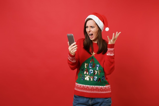 Zirytowany zły młoda dziewczyna Santa w Boże Narodzenie kapelusz krzyczy, rozkładając ręce trzymając telefon komórkowy na białym tle na czerwonym tle. Szczęśliwego nowego roku 2019 celebracja party wakacje koncepcja. Makieta miejsca na kopię.