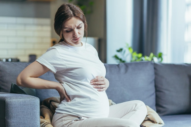 Złe samopoczucie Kobieta w ciąży trzymająca się za brzuch odczuwająca dyskomfort ból Siedząca na kanapie w domowych ubraniach