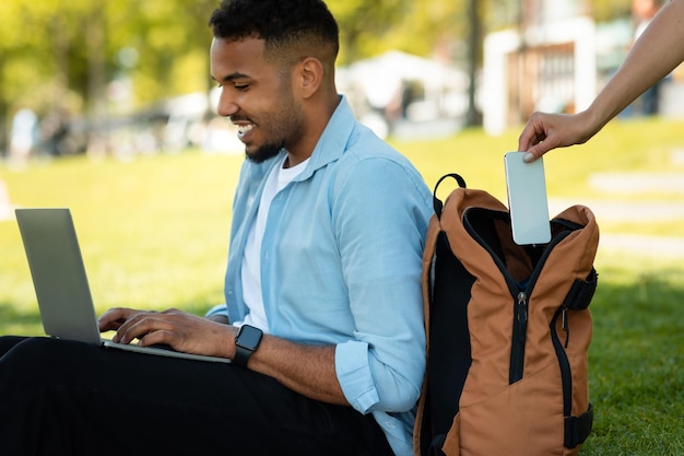 Złodziej kieszonkowy kradnący telefon z plecaka mężczyzny, podczas gdy Afroamerykanin pracuje online na laptopie w parku