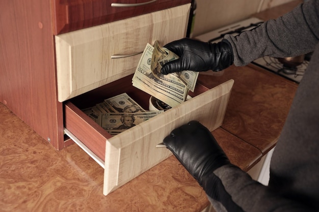 Zdjęcie złodziej w czarnym stroju i rękawiczkach widzi na otwartej półce w kuchni złodziej wyjmuje z półki banknoty dolarowe