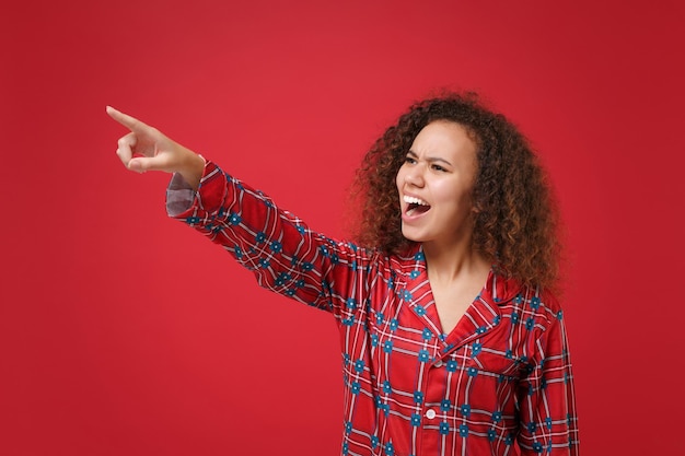 Zły młoda african american dziewczyna w piżamie homewear pozowanie, odpoczynek w domu na białym tle na czerwonym tle. Zrelaksować się koncepcja stylu życia w dobrym nastroju. Makieta miejsca na kopię. Wskazując palec wskazujący na bok, przeklinając.