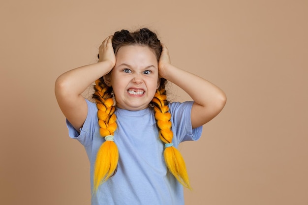 Zły obrażony zdenerwowany kaukaski mała dziewczynka trzymając głowę szczerząc zęby patrząc na kamery posiadające warkocze kanekalon żółty kolor w niebieskiej koszulce na beżowym tle