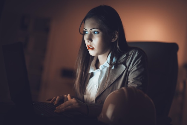 Zmartwiona samotna młoda kobieta patrząca na oświetlony ekran laptopa w nocy w domu. Jest zmęczona, bo pracuje do późna.