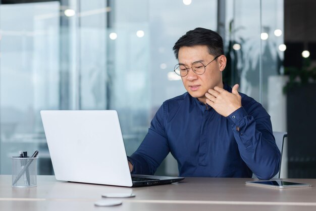 Zmartwiony i zmęczony młody azjatycki mężczyzna pracujący w biurze na laptopie czuje się źle, a temperatura jest gorąca