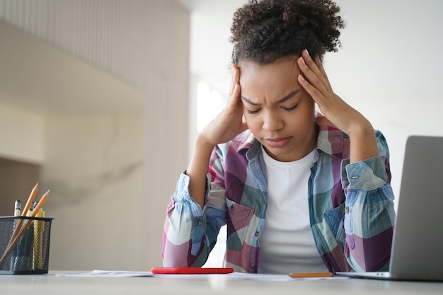 Zdjęcie zmęczona afroamerykańska nastolatka cierpi na ból głowy, odrabiając pracę domową na laptopie trudne e-learning
