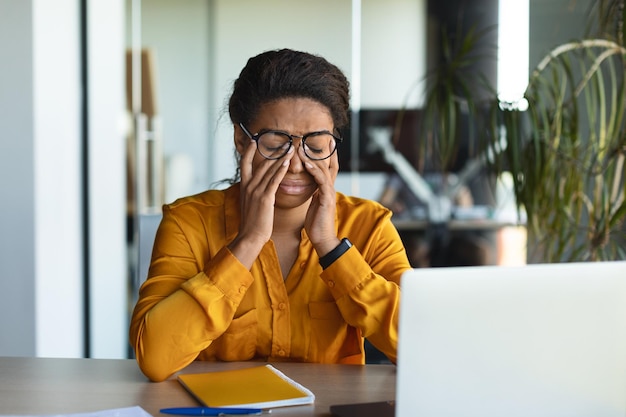 Zmęczona czarna bizneswoman cierpiąca na ból głowy i zmęczenie przecierająca oczy w miejscu pracy z laptopem we wnętrzu biura