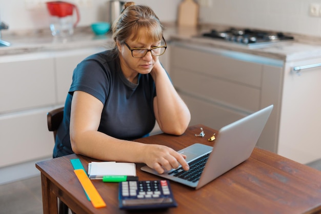 Zmęczona dojrzała blond kobieta freelancer pracująca w domu przy użyciu laptopa siedzącego w kuchni
