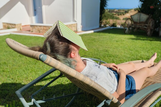 Zmęczona Dziewczyna śpi Z Książką Na Twarzy. Teen Dziewczyna Leżąc Na Leżaku Ogrodowym Na Trawniku W Pobliżu Domu, Słoneczny Letni Dzień