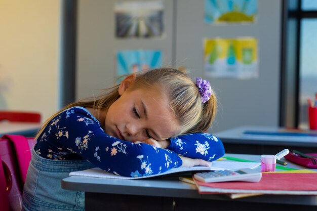 Zdjęcie zmęczona kaukaska uczennica siedząca przy biurku i leżąca na książkach w klasie w szkole podstawowej