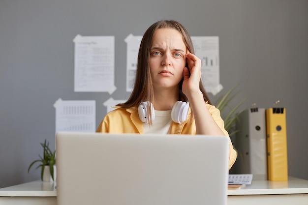 Zmęczona kobieta siedząca przed komputerem, czująca zmęczenie, cierpiąca na ból głowy, spędzająca długie godziny na pracy online, trzymając laptopa masując świątynię, patrząc na kamerę