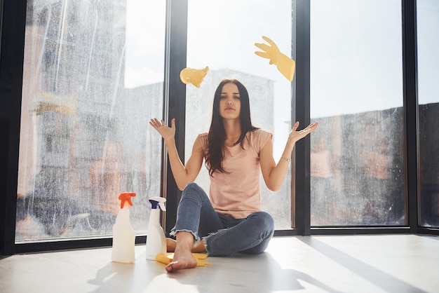Zmęczona kobieta siedzi na parapecie z butelkami sprayu do czyszczenia i wyrzuca rękawiczki w powietrze