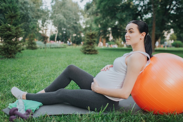 Zmęczona młoda kobieta w ciąży siedzieć na kolega z jogi w Arce. Pochyla się do pomarańczowej piłki fitness i trzyma ręce na brzuchu. Model patrzy w lewo.