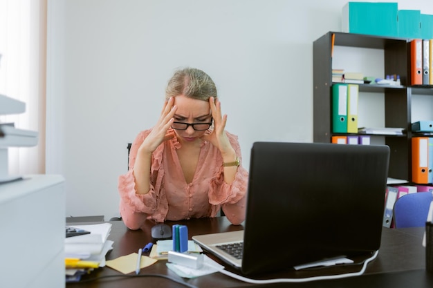 Zmęczona pracownica biurowa trzymająca się za głowę siedząc w swoim miejscu pracy w biurze