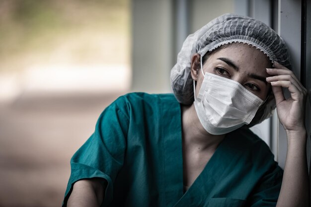 Zmęczona przygnębiona kobieta azjatycka pielęgniarka szorująca nosi maskę na twarz w niebieskim mundurze siedzi na podłodze w szpitaluMłoda lekarka zestresowana ciężką pracą