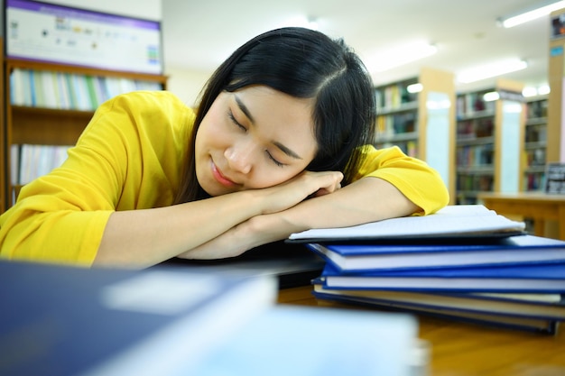 Zmęczona studentka śpi na stosie książek w bibliotece z zamazanym regałem na tle