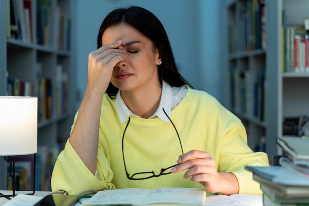 Zmęczona, zmęczona studentka przepracowuje się nocą nad projektem badawczym w bibliotece Kobieta rozmyślająca nad problemami z wypracowaniem, siedząca przy stole z dużą ilością otwartych książek, trzymająca się w głowie, czując ból głowy