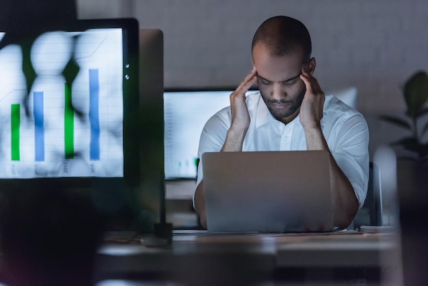 Zmęczony afroamerykański biznesmen masuje sobie skronie podczas pracy z laptopem w biurze późno w nocy