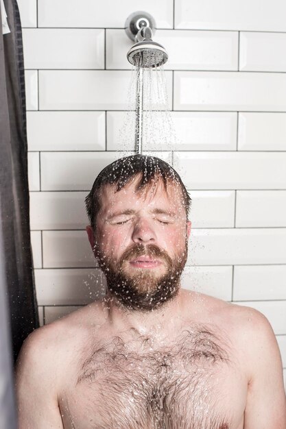 Zmęczony brodaty mężczyzna relaksuje się w łazience stojąc pod prysznicem pod bieżącą ciepłą wodą.