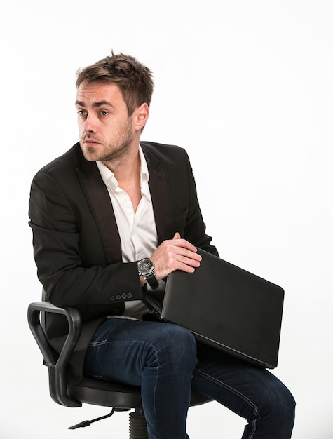 Zmęczony kierownik siedzący na krześle obejmuje laptopa, abyś nie mógł zobaczyć, nad czym pracował ze strachem, patrząc na prawo. Studio, białe tło.