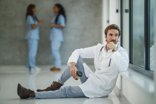 Zmęczony lekarz siedzący na podłodze i zamyślony wyglądający przez okno podczas krótkiej przerwy na kawę w korytarzu szpitala podczas pandemii Covid-19.