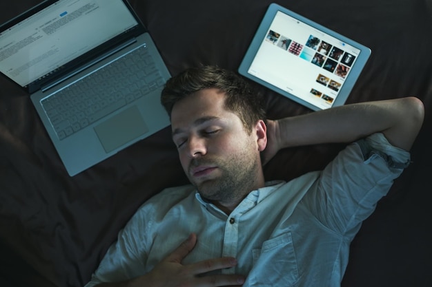 Zmęczony młody człowiek rasy kaukaskiej w białej koszuli śpiący i trzymający jedną rękę nad głową leżący na łóżku w pobliżu laptopa