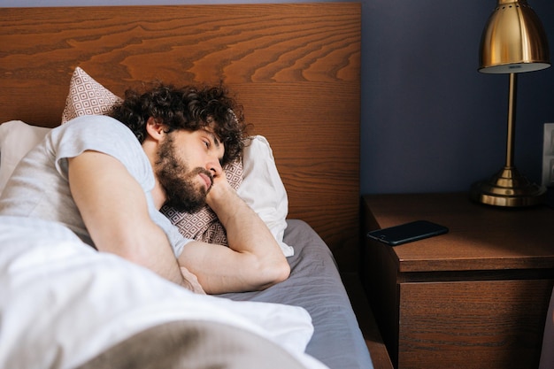 Zdjęcie zmęczony przystojny brodaty młody mężczyzna śpi z otwartymi oczami spokojnie leżąc na plecach w dużym wygodnym podwójnym łóżku pod białym kocem