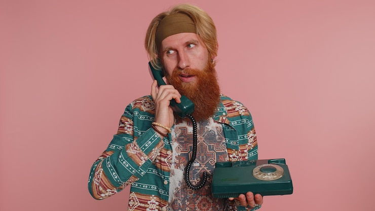 Zdjęcie zmęczony, szalony hippie rozmawiający przez przewód, vintage telefon z lat osiemdziesiątych, robiący głupie miny.