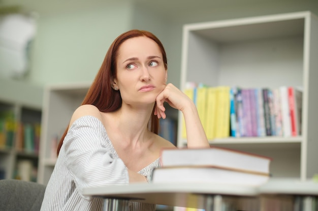 Zmęczony wygląd. Smutna młoda dość długowłosa kobieta w lekkiej bluzce siedzi przy stole z książkami w bibliotece, patrząc na bok