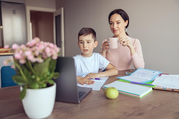 Zmieszany mały nastoletni chłopiec odrabiający pracę domową online w laptopie matki ze sprawdzaniem kawy