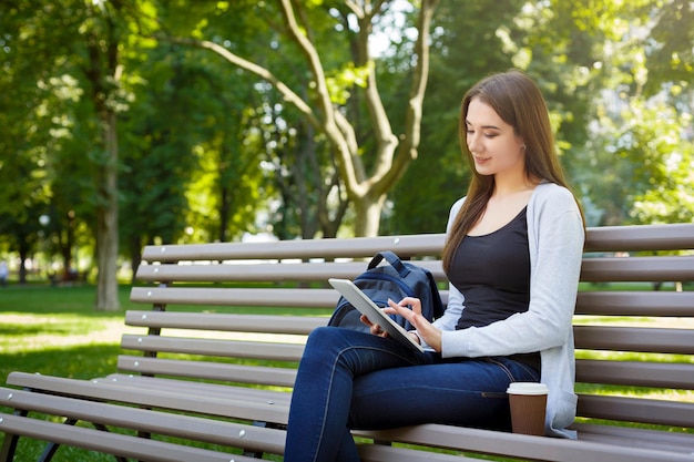 Zmotywowany azjatycki freelancer w parku. Młoda brunetka dziewczyna na ławce, picia kawy i pracy z tabletem online. Styl życia, technologia i koncepcja pracy zdalnej