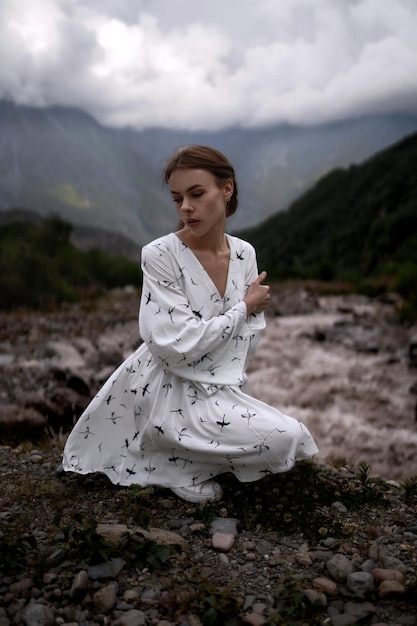 Zmysłowa elegancka dziewczyna o brązowych włosach w białej sukience siedzi w górach