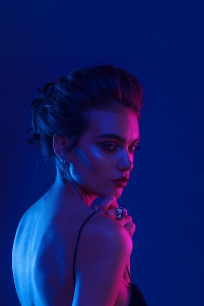 Zmysłowa młoda kobieta z odkrytymi ramionami, fryzurą i makijażem, nad ciemnofioletowymi neonami.